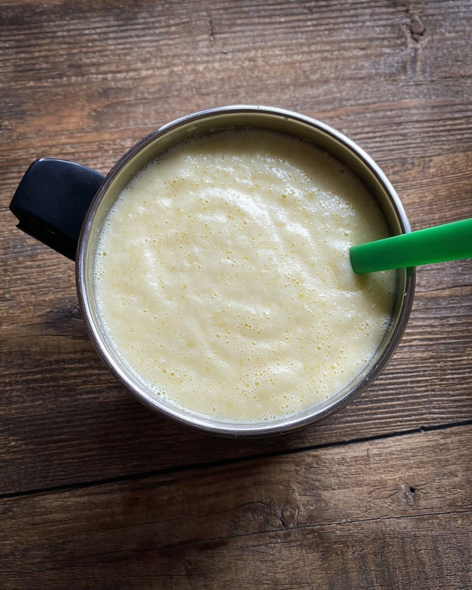 A close-up image of a metal bowl filled with a thick, light yellow creamy mixture with a slightly bubbly texture on the surface. A green spoon is partly dipped inside the mixture, resting near the right edge of the bowl. The bowl has a black handle on the left side, and it sits on a wooden surface with visible grain and texture. photo taken with an iphone --ar 4:5 --v 7