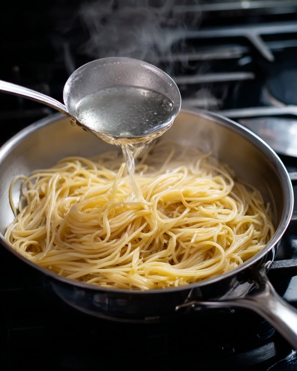 A silver pan filled with cooked spaghetti, light yellow in color, with strands loosely arranged in the pan. A shiny metal ladle holding clear hot water is being poured over the pasta, with visible steam rising around it. The pan is on a black stovetop with a small part of a silver burner visible on the right side. photo taken with an iphone --ar 4:5 --v 7