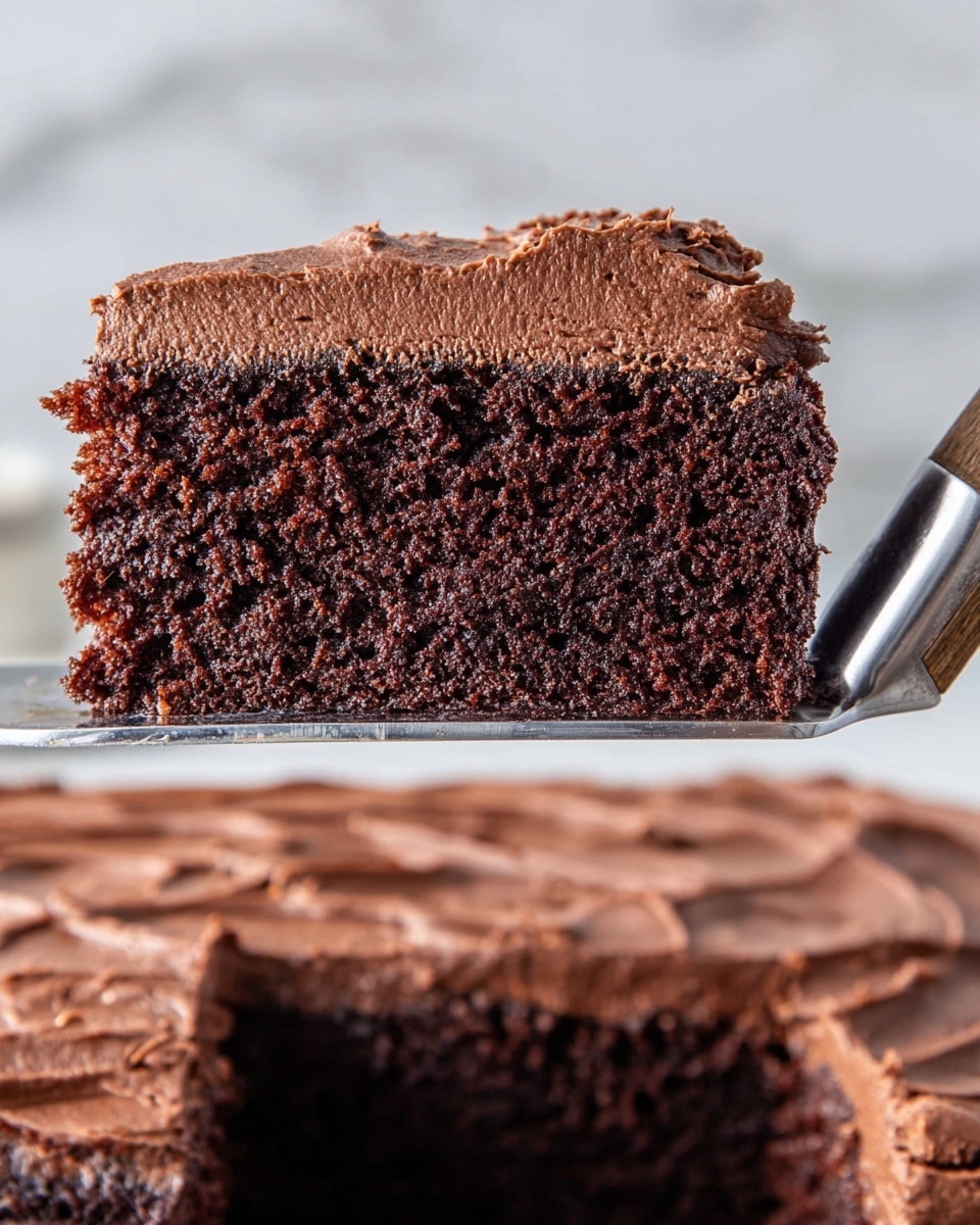 A close-up slice of chocolate cake is shown held by a metal spatula with a wooden handle. The cake has two layers: a thick, dark brown, moist-looking bottom cake layer with a soft texture, and a thick top layer of creamy, smooth chocolate frosting with a slightly rough but fluffy surface. In the background, part of the remaining frosted cake is visible, lying flat on a white marbled surface. The lighting highlights the rich, dark colors and textures of the cake. photo taken with an iphone --ar 4:5 --v 7