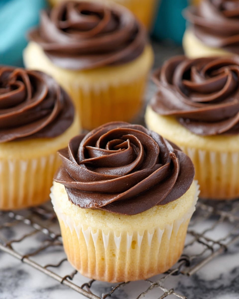 Several light yellow cupcakes with smooth, creamy dark brown chocolate frosting swirled on top in a rose pattern. Each cupcake has one layer of soft cake topped with a thick layer of shiny chocolate frosting. They sit close together on a wire rack over a white marbled texture. The focus is on the front cupcake with others softly blurred in the background. Photo taken with an iphone --ar 4:5 --v 7