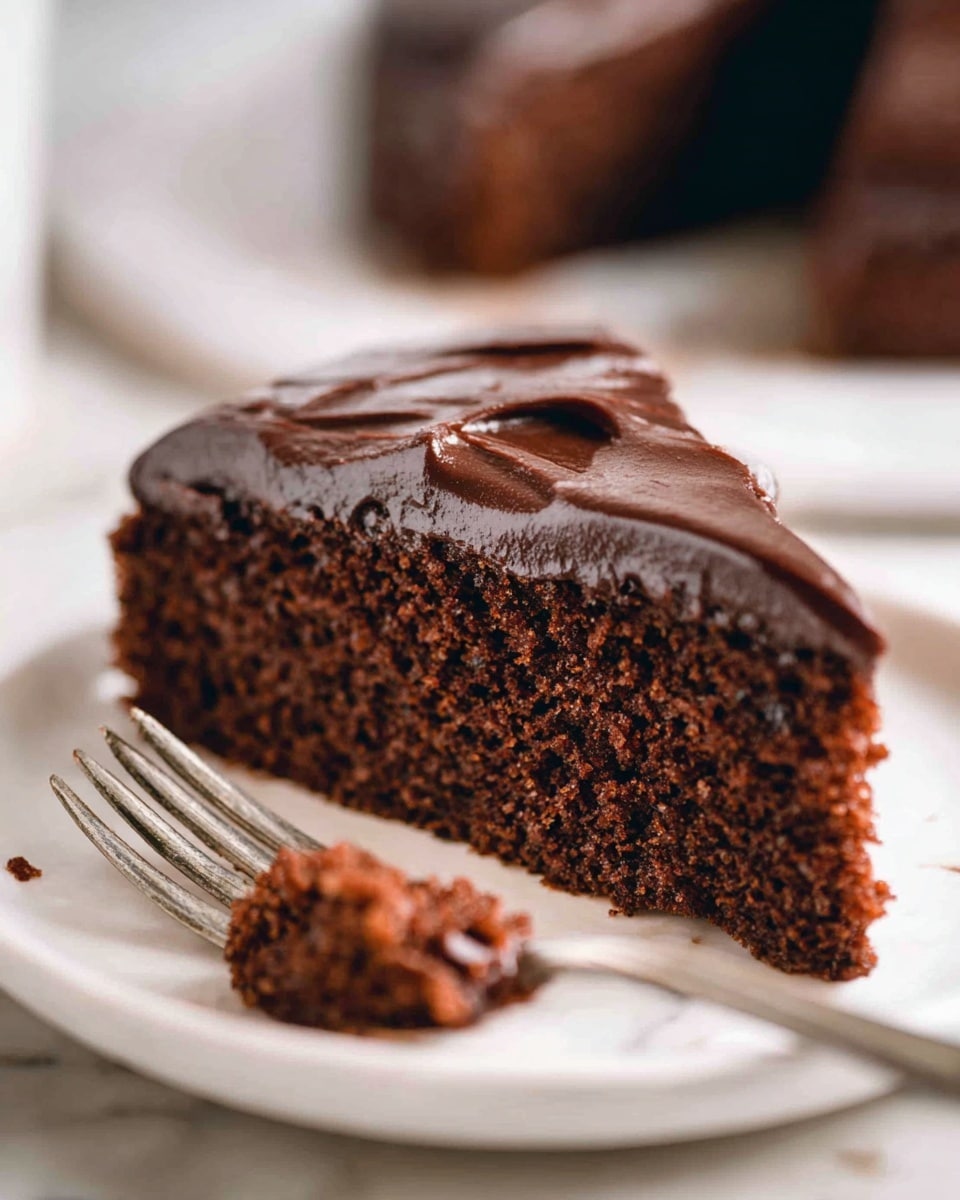 The image shows a close-up of a slice of chocolate cake with one thick layer of dark brown, moist-looking cake topped with a smooth, glossy layer of dark chocolate frosting that has visible swirl marks. The slice is placed on a white plate on a white marbled surface. In front of the slice, there is a small broken piece of cake with some crumbs scattered around. In the background, a blurred silver fork stands upright. The photo taken with an iphone --ar 4:5 --v 7