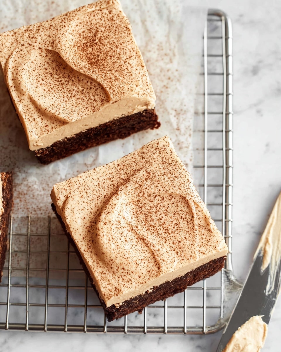 Two square-shaped dessert pieces sit on a metal cooling rack over a white marbled background. Each piece has two layers: a dark brown base with a rough texture and a thick, smooth light tan frosting layer on top. The frosting is dusted with a light sprinkle of cocoa powder, giving a delicate speckled look. The frosting is spread unevenly with visible swirls. A knife with some frosting on its blade rests on the bottom right corner on the white marbled surface. photo taken with an iphone --ar 4:5 --v 7