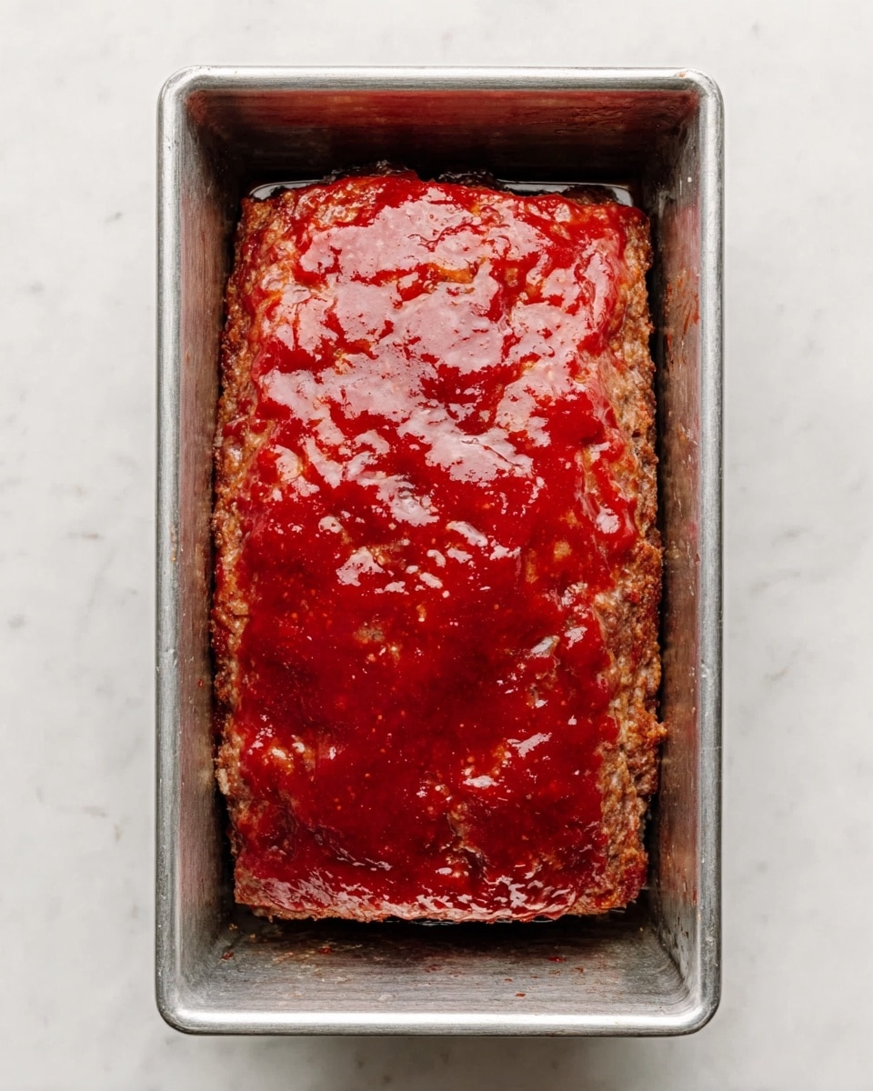 A rectangular meatloaf with a shiny red sauce covering the top sits inside a metal baking pan on a white marbled surface. The sauce is smooth and glazed, spreading evenly over the meatloaf’s textured brown surface underneath. The edges of the meatloaf are slightly rounded, and the pan fits the meatloaf snugly with clean lines and a simple look. photo taken with an iphone --ar 4:5 --v 7