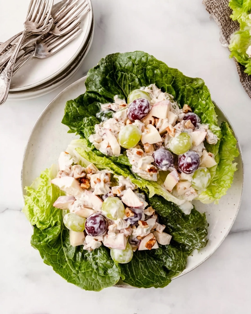A white plate holds a salad made with two large, dark green lettuce leaves forming the base layer, with a creamy mixture on top. The creamy salad contains light tan apple pieces, both green and dark purple grapes, and small bits of light brown nuts mixed with a white dressing. The plate is on a white marbled surface, with a small stack of white plates and three silver forks placed nearby. Photo taken with an iphone --ar 4:5 --v 7