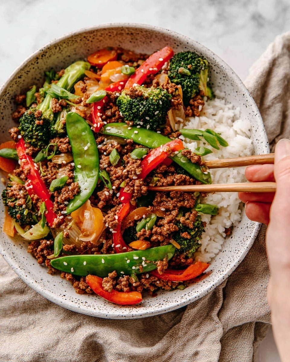 The image shows a bowl with three main layers. At the bottom, there is a layer of white rice, which is slightly visible around the edges. On top, there is a colorful stir-fry mix with several vegetables including bright green snap peas, green broccoli pieces, orange carrot slices, red bell pepper strips, and light brown cooked onions. Mixed in and covering much of the vegetables is a layer of finely cooked ground beef, brown and slightly crumbly in texture. The dish is garnished with small white sesame seeds and chopped green onion pieces, adding extra color and texture. A woman's hand holding chopsticks is picking up some of the food from the right side of the bowl. The bowl is white with some black speckles around the rim, and it sits on a beige cloth over a white marbled surface. photo taken with an iphone --ar 4:5 --v 7