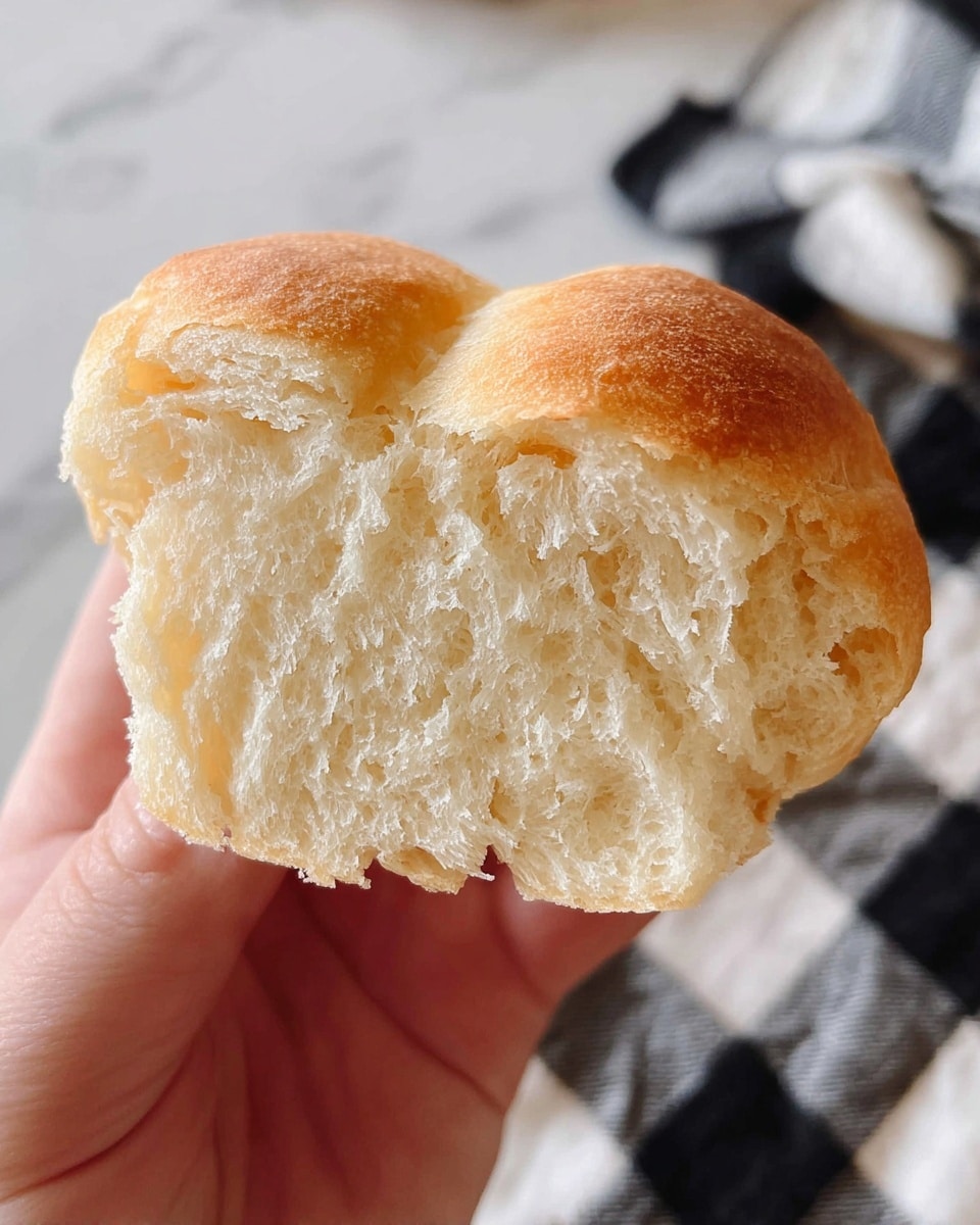 A close-up view of a piece of soft, light-colored bread held by a woman's hand. The bread shows a fluffy inner texture with small air pockets visible from a bite taken on one side. The outside crust is thin and pale golden with a smooth surface. The background features a black and white checkered cloth on a white marbled surface. photo taken with an iphone --ar 4:5 --v 7