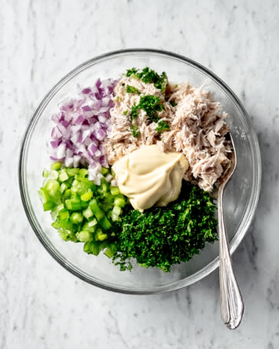 A clear glass bowl on a white marbled surface holds five separate ingredients arranged in groups inside: finely chopped red onions on the top left, chopped green celery at the bottom left, shredded light brown tuna in the center, creamy light beige mayonnaise on the top center, and chopped green herbs like parsley on the right side. A silver spoon rests beside the bowl on the right. The scene is bright and clean with natural lighting, showing fresh ingredients. photo taken with an iphone --ar 4:5 --v 7