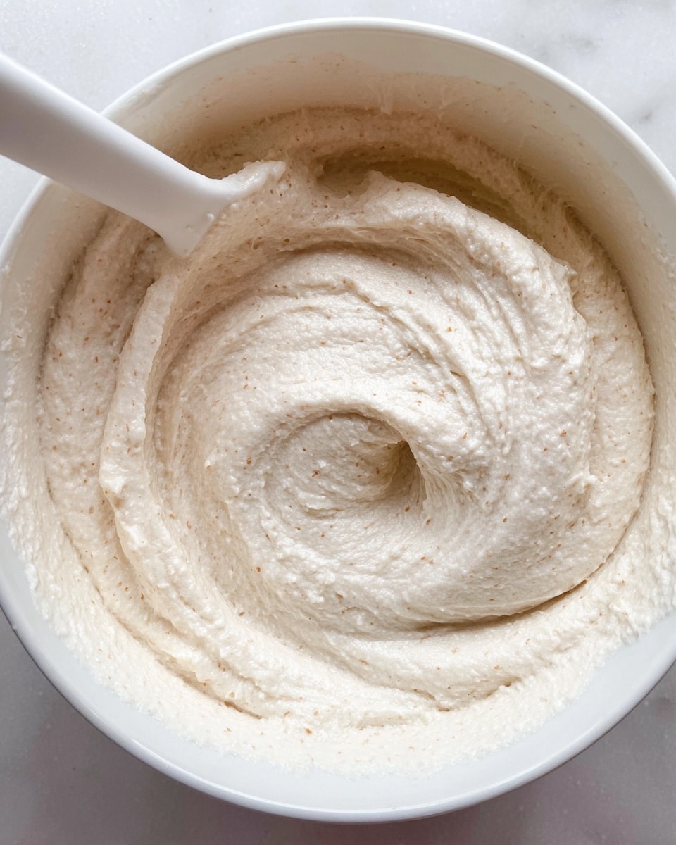 A close-up view of a thick, creamy paste inside a white bowl, showing a smooth but slightly grainy texture with small bumps throughout. The paste is light cream in color with faint beige specks, swirled in a circular motion with a white spatula partly visible at the top left corner, creating ridges and folds in the surface. The bowl is placed on a white marbled surface. photo taken with an iphone --ar 4:5 --v 7