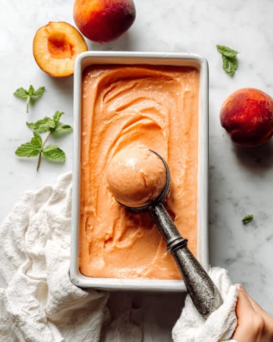 The image shows a white rectangular metal loaf pan filled with smooth, peach-colored ice cream or sorbet, having a creamy texture with soft swirls on the surface. A vintage-style metal ice cream scoop with a dark handle is lifting a rounded scoop from the middle of the pan, held by a woman's hand wrapped in a white cloth. Around the pan are two ripe peaches and some small green mint leaves scattered on a white marbled surface, adding fresh color and contrast to the scene. The lighting is soft and natural, highlighting the creamy texture of the frozen dessert and the warm tones of the peaches. photo taken with an iphone --ar 4:5 --v 7