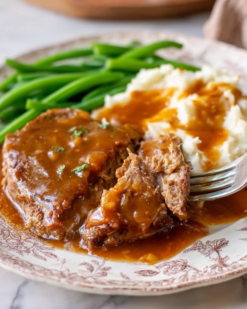 The dish shows two dark brown meat patties covered in a thick brown gravy sauce with small green herb pieces sprinkled on top. To the right, there is a mound of creamy white mashed potatoes with some gravy pouring over them. On the left side of the white plate, there is a pile of bright green cooked green beans, smooth and elongated. The plate has a detailed light gray pattern around the edges and is placed on a white marbled surface. A metal fork is visible in the top background near the mashed potatoes. photo taken with an iphone --ar 4:5 --v 7