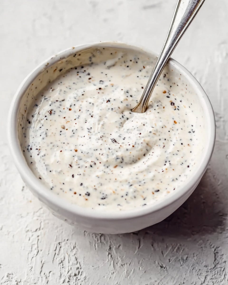 A close-up of a white bowl filled with a creamy white sauce that has small black, brown, and beige specks mixed throughout; the sauce appears thick and slightly textured. Inside the bowl, a silver spoon with a simple design is partially immersed in the sauce, standing upright. The bowl rests on a white marbled textured surface, giving a clean and fresh look. Photo taken with an iphone --ar 4:5 --v 7