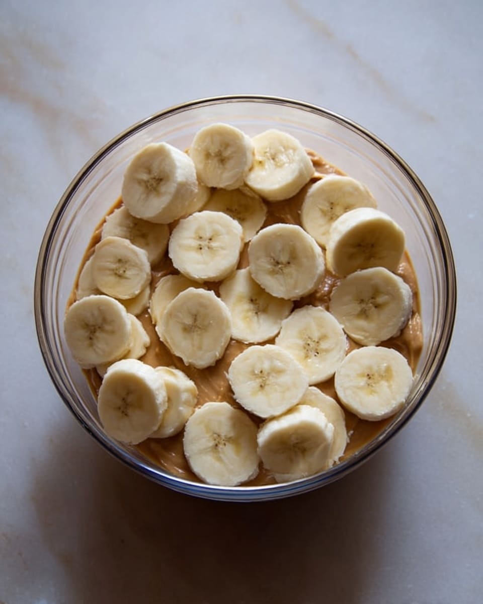 The image shows several round slices of banana arranged on top of a layer of smooth peanut butter inside a clear glass bowl. The banana slices are light yellow with a soft texture, layered evenly across the peanut butter, which is a rich brownish-tan color and fills the bottom of the bowl. The bowl sits on a white marbled surface, and the inside of the bowl reflects some light, giving it a clear and shiny look. photo taken with an iphone --ar 4:5 --v 7