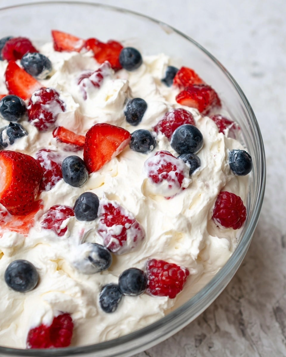 A clear glass bowl filled with a creamy white mixture that holds chunks of white fluffy texture combined with bright red strawberry slices, dark blue blueberries, and a few red raspberries scattered evenly throughout. The mixture looks soft and thick, with the fruit pieces peeking through in all places. The bowl sits on a light brown fabric with a white marbled background under it. photo taken with an iphone --ar 4:5 --v 7