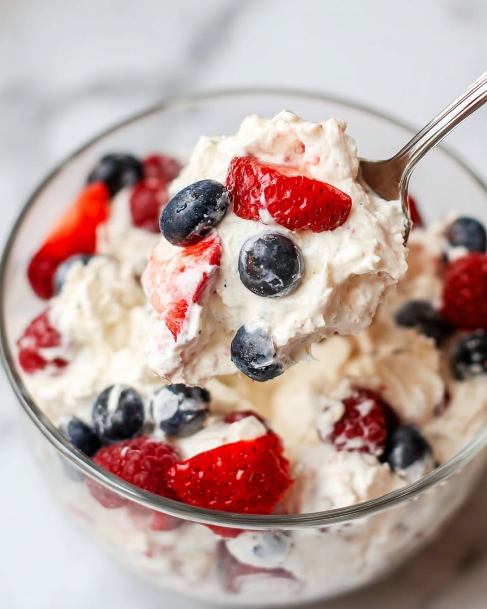 A clear glass bowl is filled with three layers: a fluffy white cream base, which is mixed with bright red raspberries, deep blue blueberries, and sliced medium red strawberries. The cream texture is soft and light, with fresh berries scattered throughout, and the spoon lifting a scoop holds a mix of white cream with a whole raspberry, a whole blueberry, and a sliced strawberry on top, showing the vibrant colors and juicy textures of the berries against the smooth cream. The scene is set on a white marbled surface photo taken with an iphone --ar 4:5 --v 7