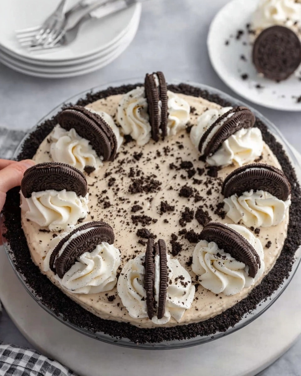 The image shows a round Oreo cookie pie with a dark, crumbly crust. On top of the crust is a smooth cream layer sprinkled with tiny Oreo crumbs. Around the edge, there are eight swirled dollops of whipped cream, each topped with a small Oreo cookie piece sliced in half and placed upright. The pie is on a white marbled surface with a white plate and fork in the background. A woman's hand is holding a slice of the pie. photo taken with an iphone --ar 4:5 --v 7