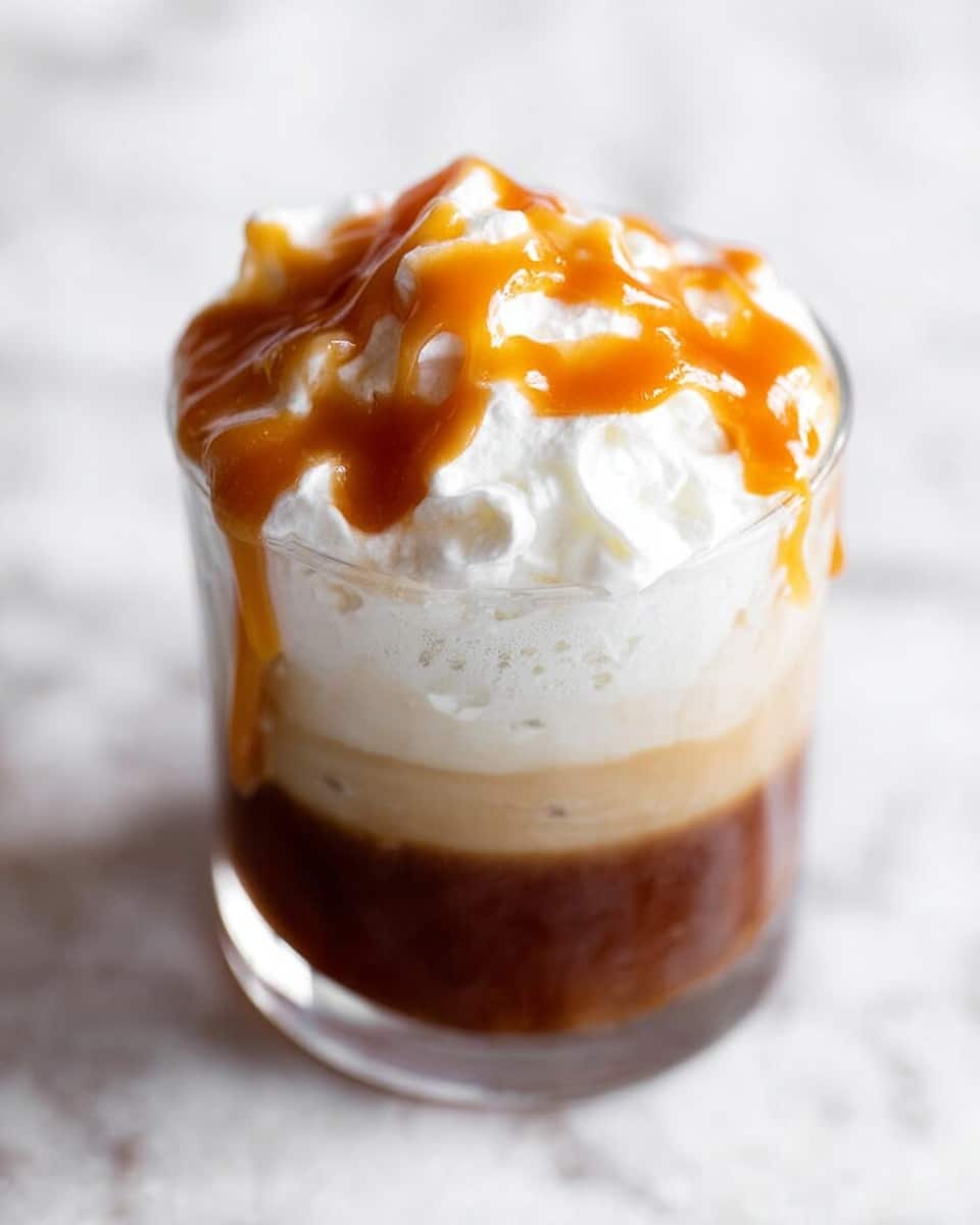 A clear glass filled with three layers: the bottom layer is dark brown liquid, the middle layer is creamy light beige, and the top layer is white whipped cream with soft peaks. On top of the whipped cream, there is a thick, shiny orange caramel sauce that slightly drips over the side of the glass. The glass sits on a white marbled textured surface in soft light. photo taken with an iphone --ar 4:5 --v 7