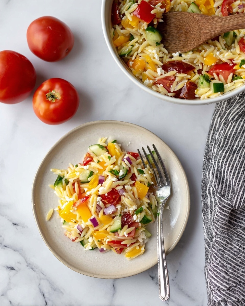 A small white plate sits on a white marbled surface, holding a colorful orzo pasta salad with visible layers of yellow, red, and orange chopped bell peppers, green cucumber pieces, red onion bits, small chunks of red tomatoes, and crumbled white feta cheese mixed through pale yellow orzo pasta. A silver fork rests angled on the plate. To the right of the plate is a striped grey and white cloth. In the top right corner, a white bowl filled with the same orzo salad shows a wooden spoon resting inside it. Two whole red tomatoes lie to the left of the bowl, all set against the white marbled background. Photo taken with an iphone --ar 4:5 --v 7