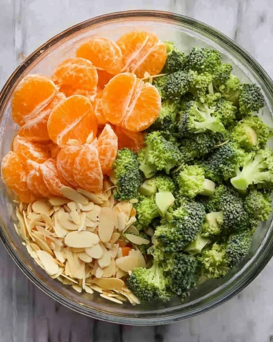 The image shows a clear glass bowl filled with three layers of fresh ingredients. On the left side, there are bright orange tangerine segments with a smooth, shiny texture. Next to them, on the right side, are small green broccoli florets with a rough, bumpy texture. Below the broccoli and tangerines, there are pale yellow sliced almonds with a thin and flat texture spread out evenly. The bowl sits on a white marbled surface. photo taken with an iphone --ar 4:5 --v 7