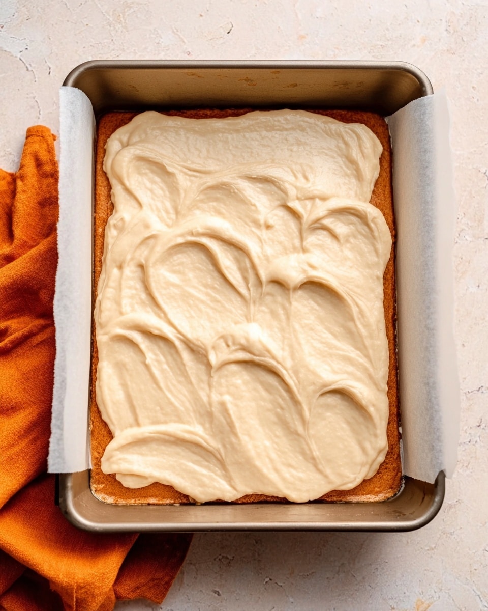 A rectangular baking tray lined with white parchment paper holds a layer of smooth, pale tan batter spread unevenly across the surface, showing some soft swirls and rounded edges. The batter covers the tray fully but is not perfectly even, with gentle peaks and slight lumps. The tray sits on a white marbled surface with an orange cloth partially visible to the side. photo taken with an iphone --ar 4:5 --v 7