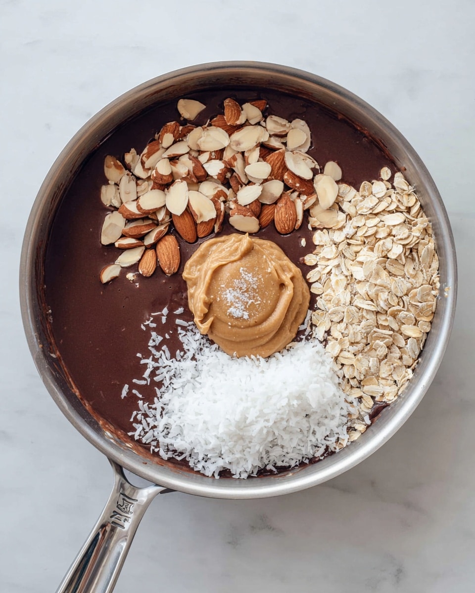 A top view of a stainless steel pan filled with a dark brown smooth base, likely chocolate or similar. On top of this base, four distinct toppings are arranged in layers: the top left portion is covered with toasted almond slices showing shades of light brown and tan with a slight shine; the center right features light beige rolled oats scattered loosely; below the oats on the right is a fluffy white shredded coconut layer with a rough texture; near the bottom center there is a scoop of creamy light brown peanut butter topped with a small pinch of white salt. The pan is placed on a white marbled surface. photo taken with an iphone --ar 4:5 --v 7