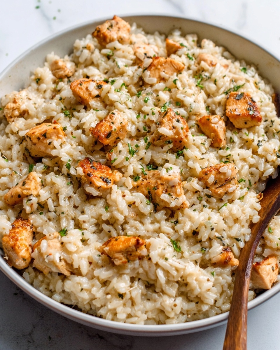 A close-up of a white bowl filled with a creamy risotto dish, featuring small pieces of golden-brown cooked chicken scattered evenly throughout the creamy light beige rice. The risotto looks slightly glossy and is garnished with small bits of green herbs on top. The bowl sits on a white marbled surface with a wooden spoon handle visible on the right side of the image. The texture of the rice is soft and slightly sticky, and the chicken pieces have a slightly crispy outside. photo taken with an iphone --ar 4:5 --v 7