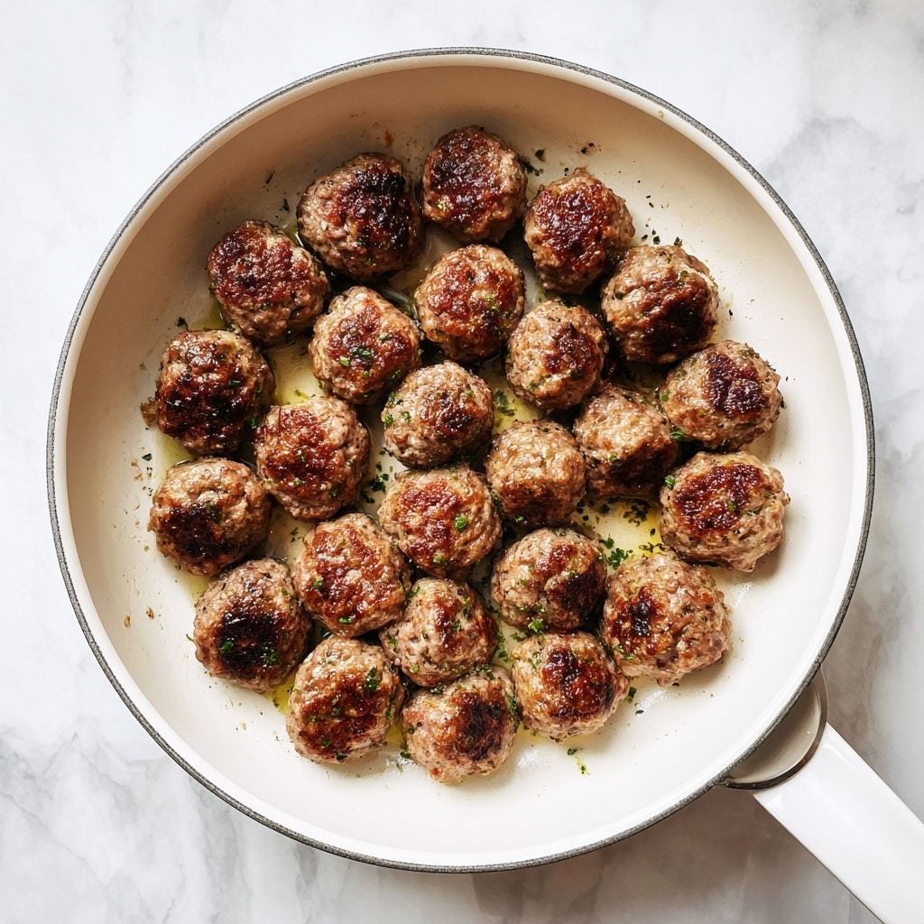 A white bowl filled with small round meatballs, each browned on top with a slightly crispy texture. The meatballs sit on a bed of creamy, light beige mashed potatoes that have a smooth surface with some black pepper sprinkled on top. The bowl is placed on a white marbled surface. Photo taken with an iphone --ar 4:5 --v 7