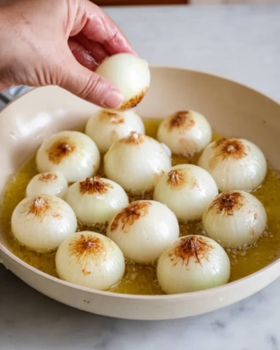 A white pan filled with about fifteen small, round white onions with slightly brown tops cooking in light golden oil. A woman's hand is gently placing one more onion into the pan from the top left corner. The cooked onions have a smooth, soft texture with some browned spots. The background is a white marbled surface. Photo taken with an iphone --ar 4:5 --v 7
