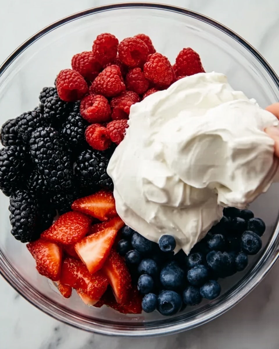 A clear glass bowl holds four sections arranged side by side: bright red raspberries in the top left, shiny black blackberries in the bottom left, deep blue blueberries in the bottom right, and fresh sliced red strawberries in the top right. A large dollop of thick white whipped cream is being added by a woman's hand, hovering just above the berries. The bowl sits on a white marbled surface. The photo taken with an iphone --ar 4:5 --v 7