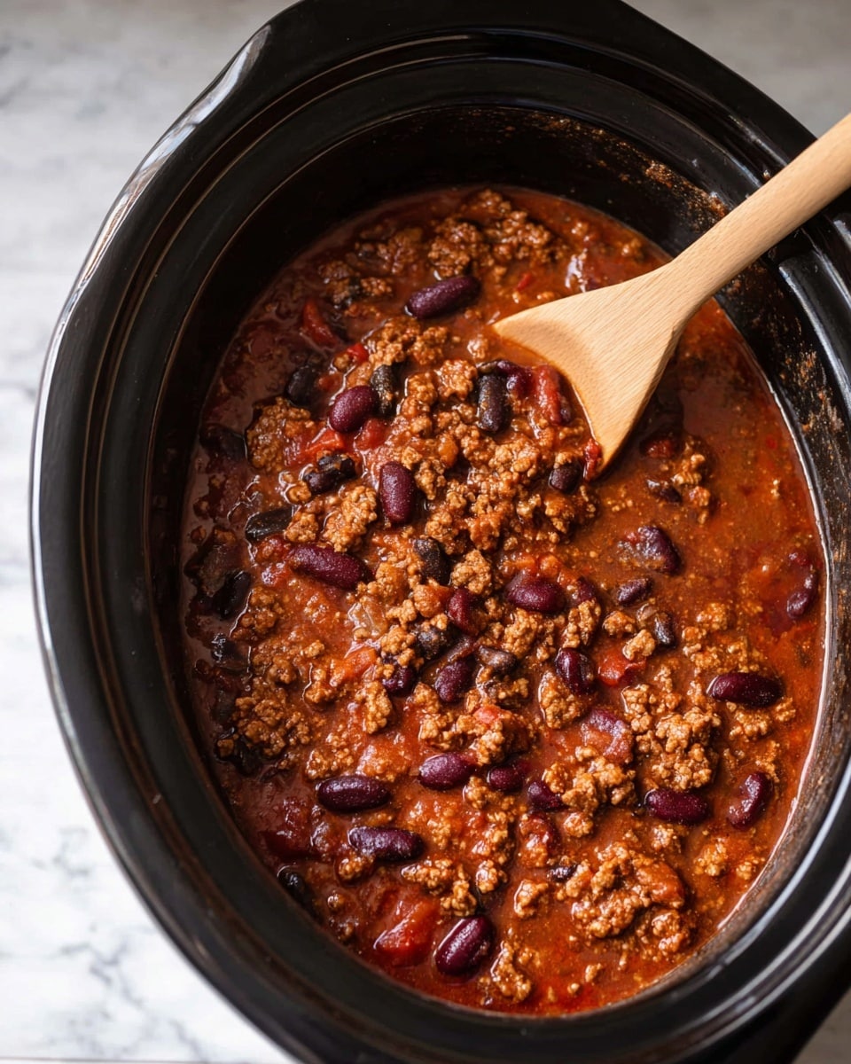 A close-up of a black slow cooker filled with thick chili showing three layers: a reddish-brown sauce forming the base layer, a middle layer of cooked ground meat in clumps with a crumbly texture, and scattered dark red and black beans as the top layer. A light brown wooden spoon rests inside the slow cooker on the right side, partially covered by the chili. The slow cooker sits on a white marbled surface. Photo taken with an iphone --ar 4:5 --v 7