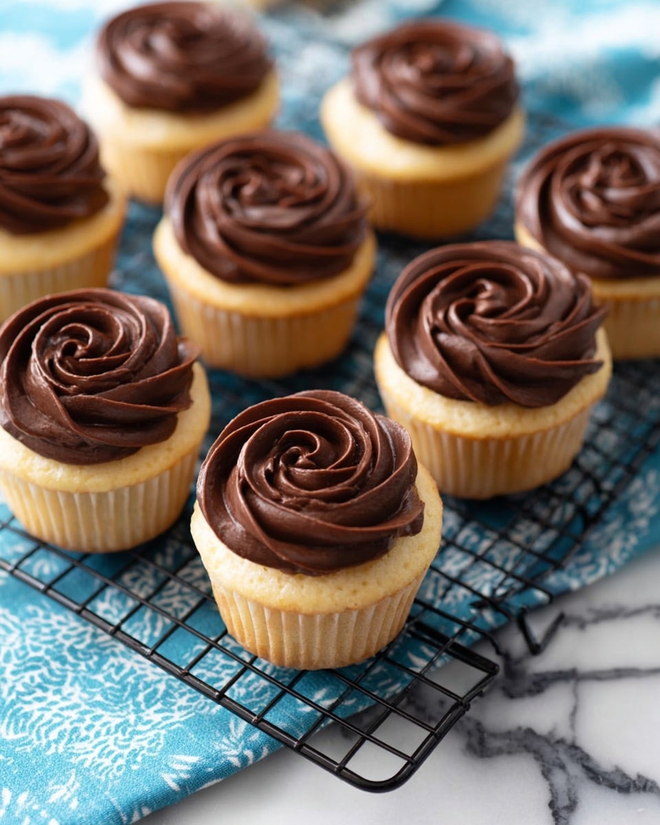 There are seven vanilla cupcakes arranged on a black cooling rack placed over a white marbled surface with a blue and white cloth beneath. Each cupcake has a light golden base with a visible texture of soft cake and is topped with a thick swirl of smooth, rich dark chocolate frosting that looks shiny and creamy. The chocolate frosting is shaped in a rose-like pattern on top of each cupcake. Photo taken with an iphone --ar 4:5 --v 7