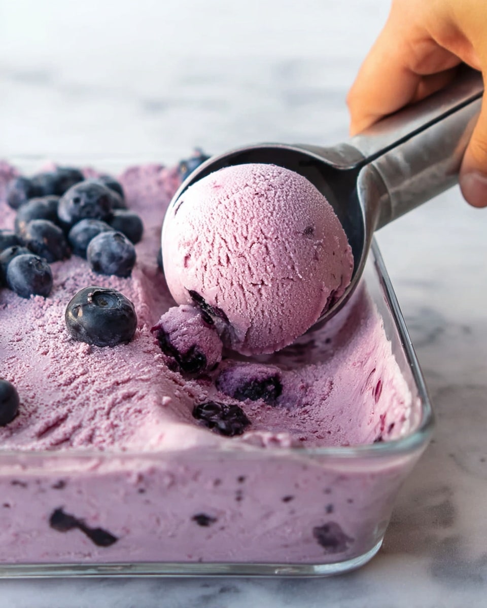 A white bowl holds three scoops of purple ice cream with small bits of brown sprinkled on top. The ice cream looks smooth and creamy. Around the bowl, fresh blueberries are scattered on a white marbled surface. Part of a graham cracker is placed near the bowl. In the background, another white bowl filled with the same purple ice cream is partly visible. A woman's hand holds a small green spoon near the bowls. photo taken with an iphone --ar 4:5 --v 7