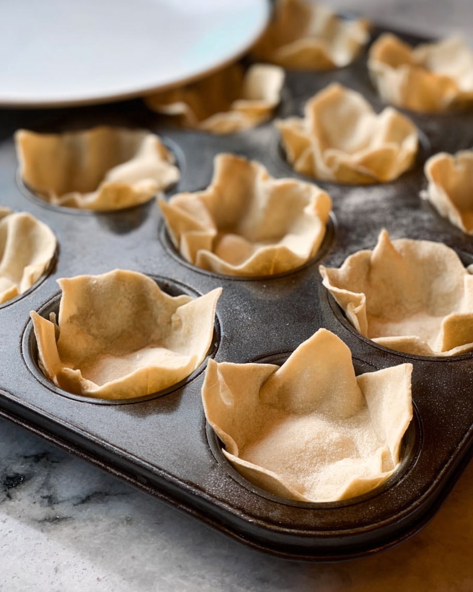 A dark metal muffin tray with 12 cups is filled with thin, beige pastry sheets pressed inside each cup. The pastry sheets have a slightly wrinkled and uneven texture, with some edges folded or bent outward, giving the cups a rustic look. The tray sits on a white marbled surface, and the background shows a white plate slightly blurred. The lighting is natural, highlighting the soft texture and pale color of the pastry. photo taken with an iphone --ar 4:5 --v 7