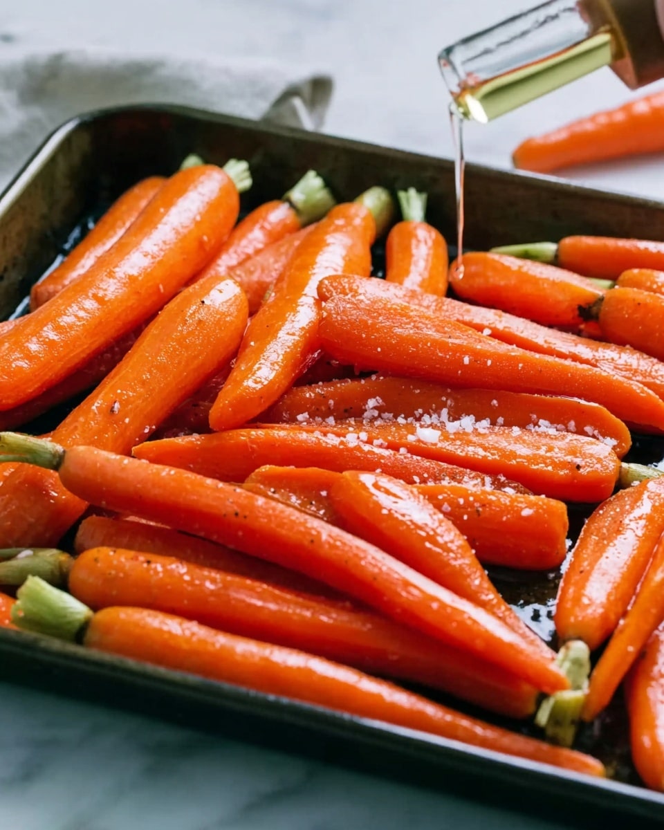 The image shows a dark metal tray filled with whole peeled carrots that are bright orange and shiny. The carrots are laid out side by side, and some have small green tops. Coarse salt and black pepper are sprinkled on top of the carrots. A dropper is held by a woman's hand, gently pouring a clear liquid over the carrots. The background is a white marbled surface. photo taken with an iphone --ar 4:5 --v 7