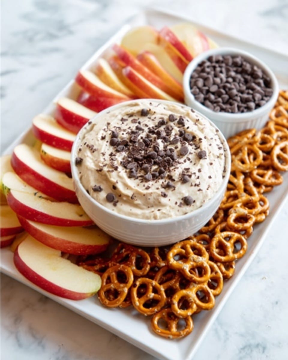 A white bowl filled with creamy dip topped with small chocolate chips sits in the center of a rectangular white plate on a white marbled surface. Arranged around the bowl are several thin, red apple slices on the top left side and a pile of small, golden-brown pretzels on the bottom right side. The dip has a smooth, light color, and the chocolate chips add a dark contrast on top. Nearby, there is a small white bowl filled with more chocolate chips. Photo taken with an iphone --ar 4:5 --v 7