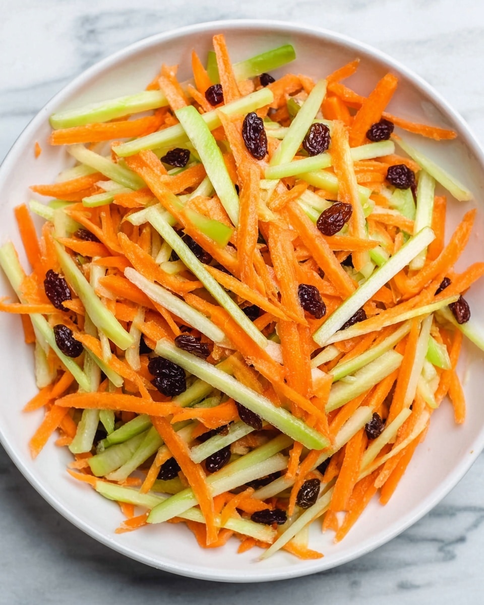 The image shows a white bowl full of a colorful salad on a white marbled surface. The salad has thin, long strips of orange carrots and light green celery mixed evenly together. There are small bits of dark brown raisins sprinkled throughout the salad, adding a contrasting color. The texture of the carrots and celery looks fresh and crunchy, with the raisins appearing soft and moist. Photo taken with an iphone --ar 4:5 --v 7