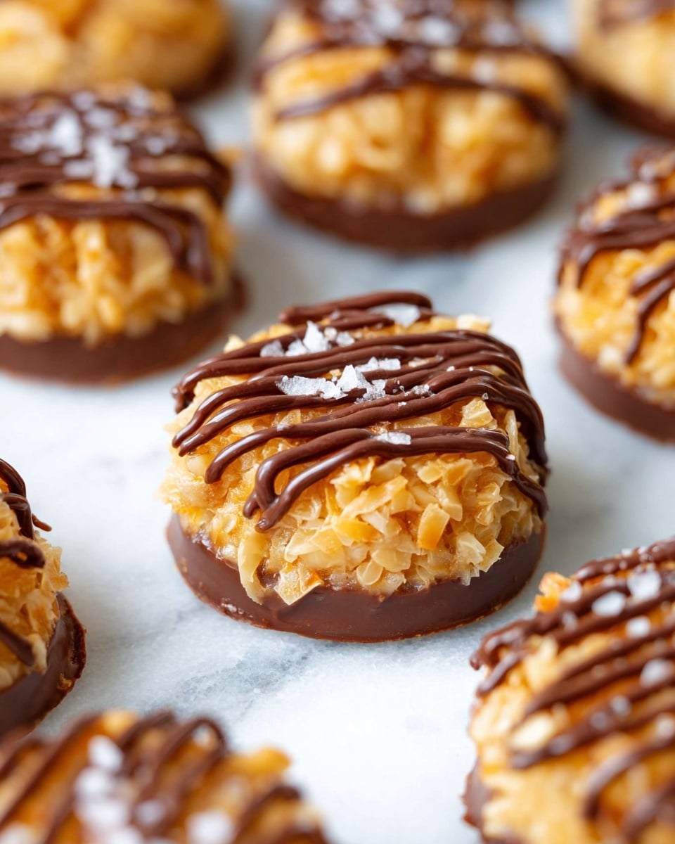 The image shows several round treats with three layers: a bottom layer of smooth, dark brown chocolate base, a thick middle layer of golden caramel mixed with shredded coconut giving a textured look, and a top layer with thin dark chocolate drizzles in lines over the caramel-coconut mix, sprinkled with small white salt flakes. The treats rest directly on a white marbled surface, and they are arranged closely together with a soft focus on the background. photo taken with an iphone --ar 4:5 --v 7