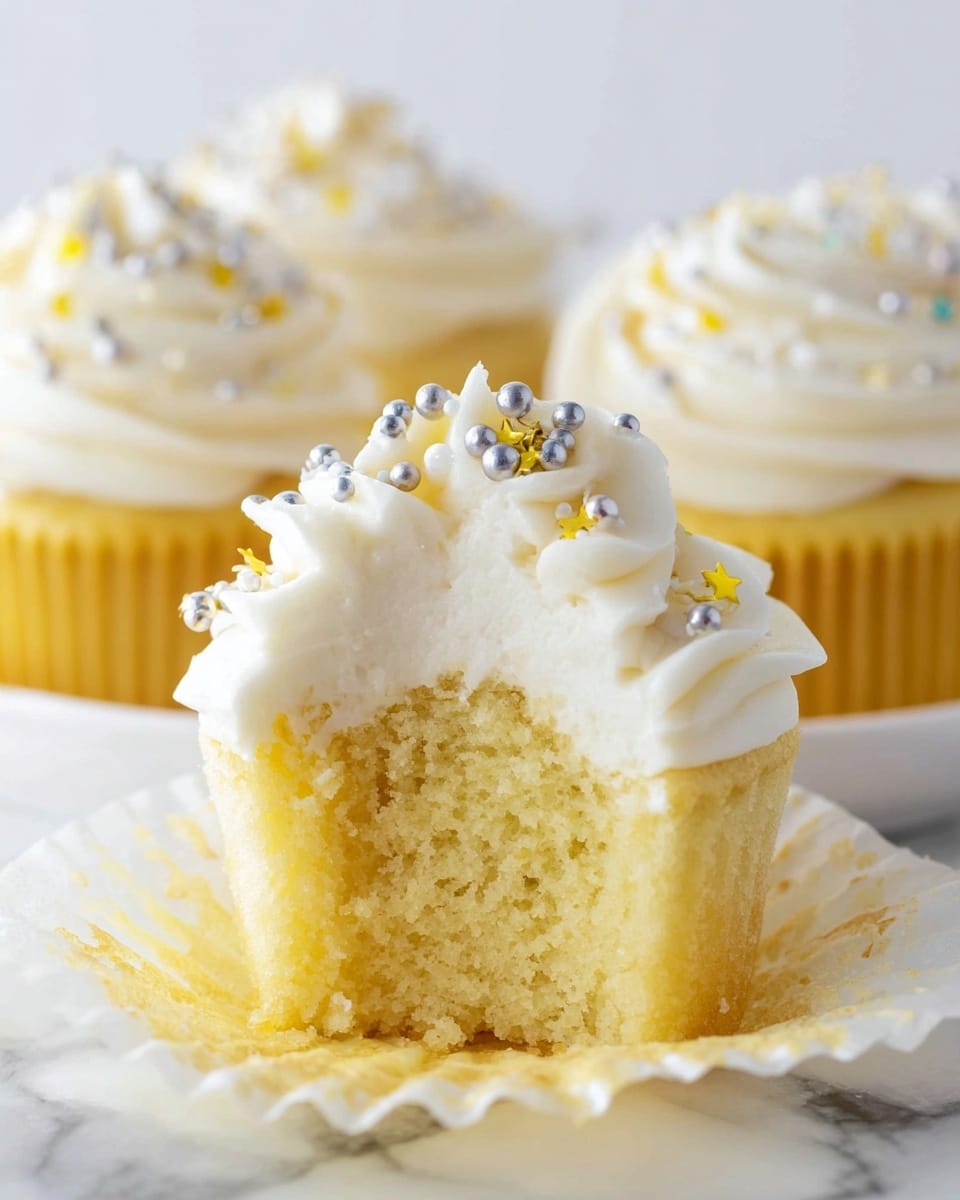 A soft yellow cupcake with a fluffy, moist inside is shown with its white cupcake liner peeled down around it, revealing the texture. On top, there is a thick layer of creamy white frosting swirled high with small, round silver, white, and tiny yellow star sprinkles scattered over it. In the background, three more similar cupcakes with the same frosting and sprinkles are visible, all resting on a white marbled surface. The focus is on the front cupcake with a bite taken out, showing the soft inside clearly. Photo taken with an iphone --ar 4:5 --v 7