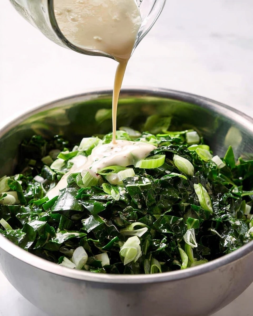 A large shiny steel bowl filled with a mix of chopped dark green leafy vegetables and small slices of light green onions. A creamy beige dressing is being poured from a clear measuring cup above, creating a small pool on top of the greens. The bowl sits on a white marbled surface, and the background is bright and clean. photo taken with an iphone --ar 4:5 --v 7