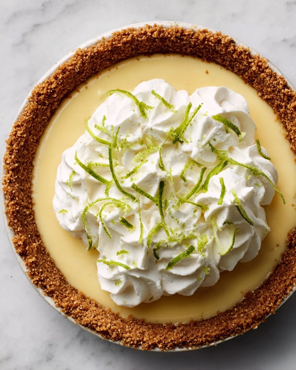 The image shows a round pie with a brown crumb crust holding a smooth, pale yellow filling. On top of the filling, there is a thick layer of white whipped cream swirled in soft peaks. Thin green strips of lime zest are sprinkled over the whipped cream, adding a fresh touch. The pie is placed on a white marbled surface. Photo taken with an iphone --ar 4:5 --v 7