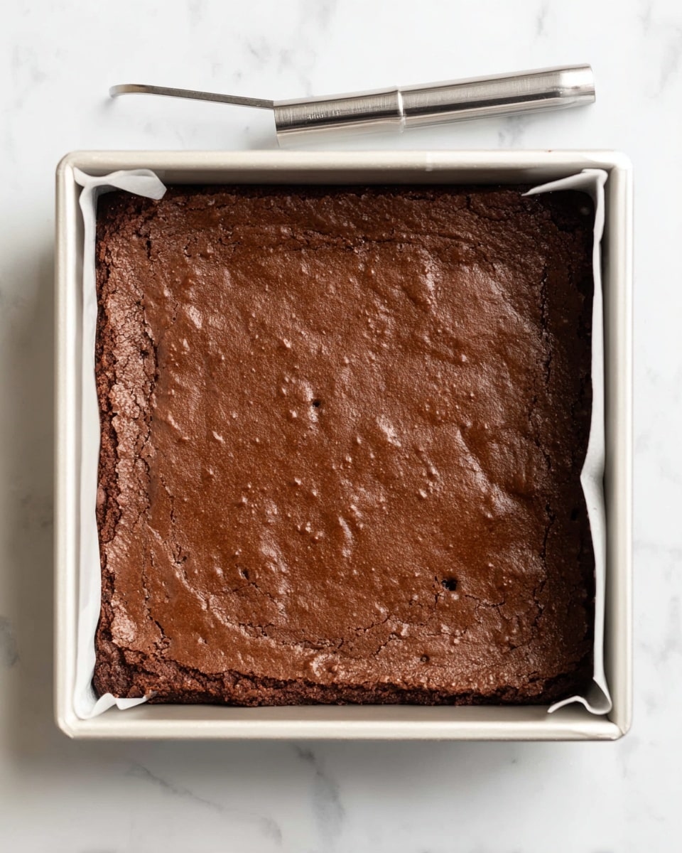 This image shows a square pan with a single thick layer of baked chocolate brownie inside. The surface of the brownie is a rich dark brown color, with a slightly shiny, cracked, and uneven texture. The edges appear a bit rough and crumbly, while the middle part is smooth with a few small bubbles and dimples. The pan has straight sides and white parchment paper liners peeking out on two opposite sides. The pan rests on a white marbled surface, and a metal cake tester stick is placed above the pan. photo taken with an iphone --ar 4:5 --v 7
