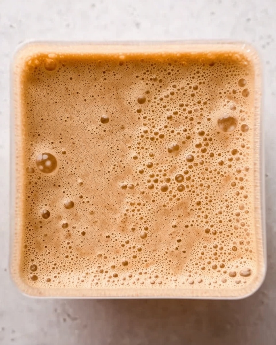 The image shows a top-down view of a square container filled with a light brown frothy liquid that has many small and medium bubbles on its surface. The container is placed on a white marbled surface, and the liquid has a smooth texture with some areas showing slightly larger bubbles. The entire focus is on the thick, foamy liquid inside the container, with no other objects visible. photo taken with an iphone --ar 4:5 --v 7