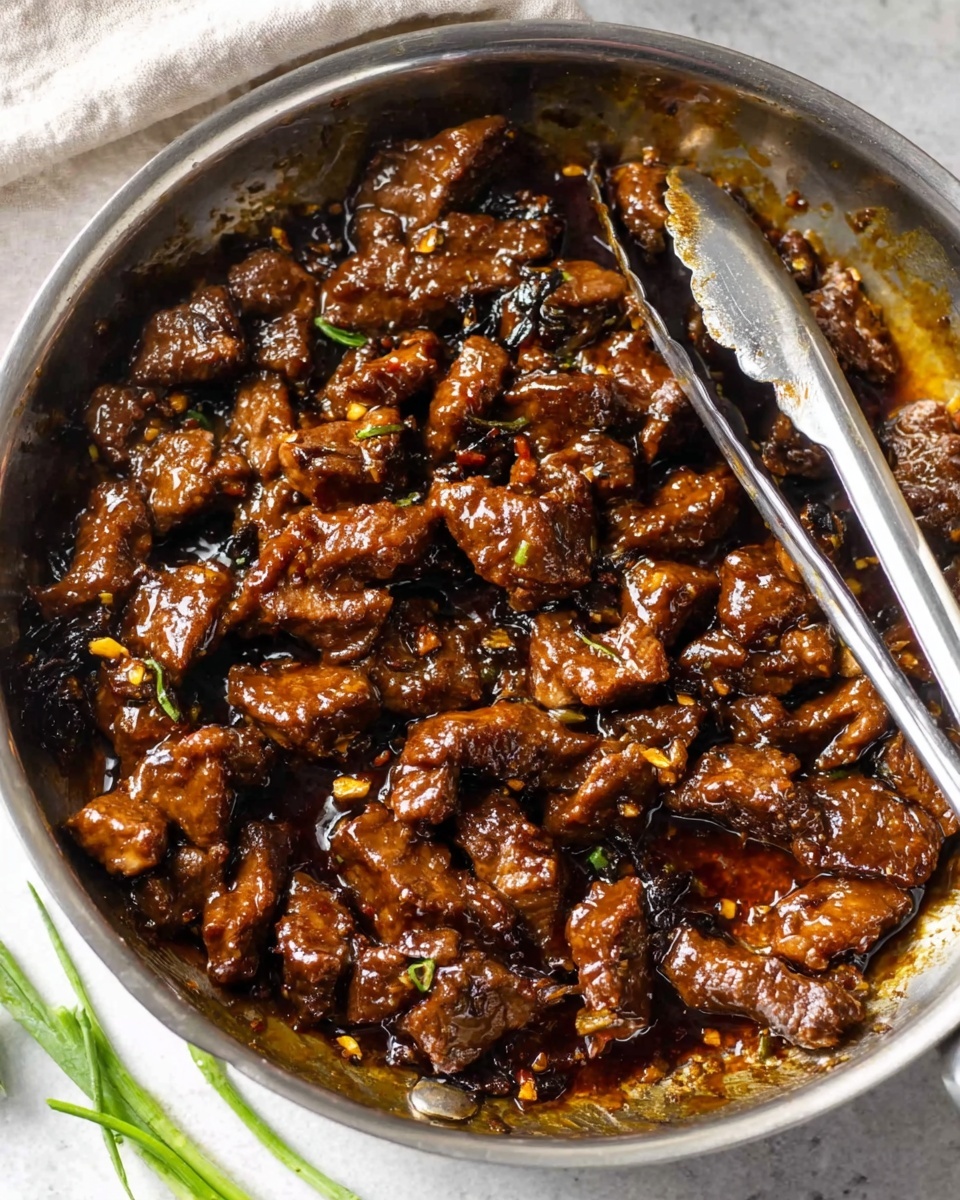 The image shows a round metal pan filled with cooked pieces of dark brown meat covered in a shiny, thick sauce. The meat pieces have a slightly uneven texture, showing they are tender and well-cooked. There are black and golden brown bits scattered throughout the dish, adding a rich look to the sauce. A pair of metal tongs rests inside the pan, with the handles extending upward and slightly outward. The pan sits on a white marbled surface, and there are a few chopped green onion pieces visible at the bottom edge of the image. The overall scene looks warm and delicious, with a rustic feel. photo taken with an iphone --ar 4:5 --v 7