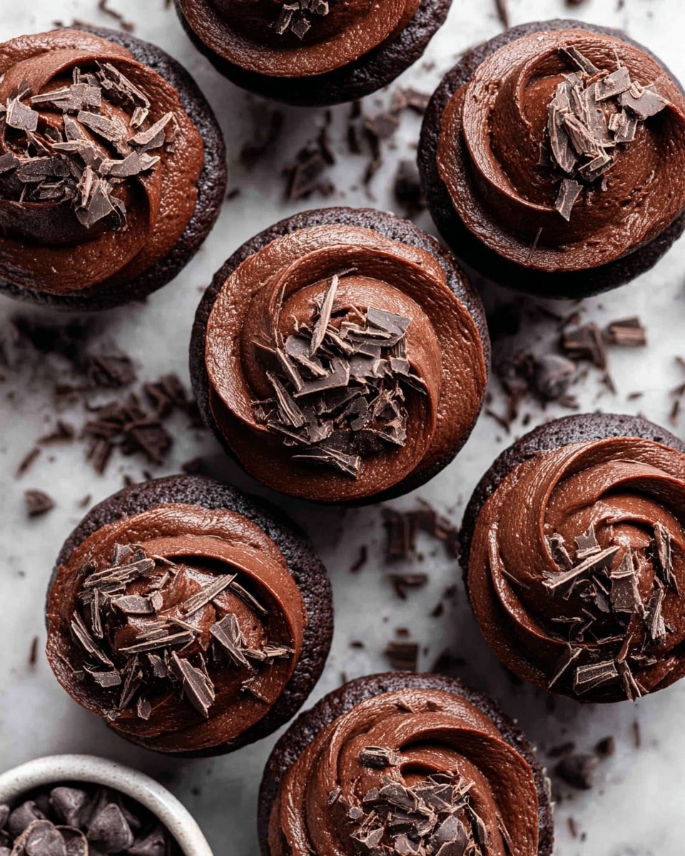 Several chocolate cupcakes are arranged closely on a white marbled surface. Each cupcake has one thick layer of rich, smooth, dark chocolate frosting spread on top in a swirled pattern. The frosting has a glossy texture and is sprinkled with small, thin shards of dark chocolate scattered evenly across the surface. Near the cupcakes, there is a white bowl filled with dark chocolate chips, adding to the chocolate theme. The overall scene is tightly zoomed in to show detail and texture of the chocolate frosting and shavings. Photo taken with an iphone --ar 4:5 --v 7