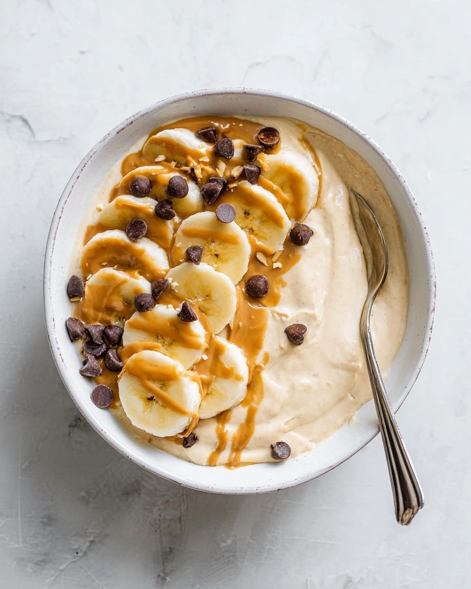 A white bowl filled with a smooth light beige base that is spread evenly across the bottom. On top, there are three thick curved rows of banana slices, light yellow with a soft texture. One row is spread across the right side, another along the left edge, partly covered with a drizzled brownish-orange peanut butter spread and scattered dark brown chocolate chips. A spoon rests inside the bowl on the left, partly under the banana slices. The bowl sits on a white marbled surface. Photo taken with an iphone --ar 4:5 --v 7