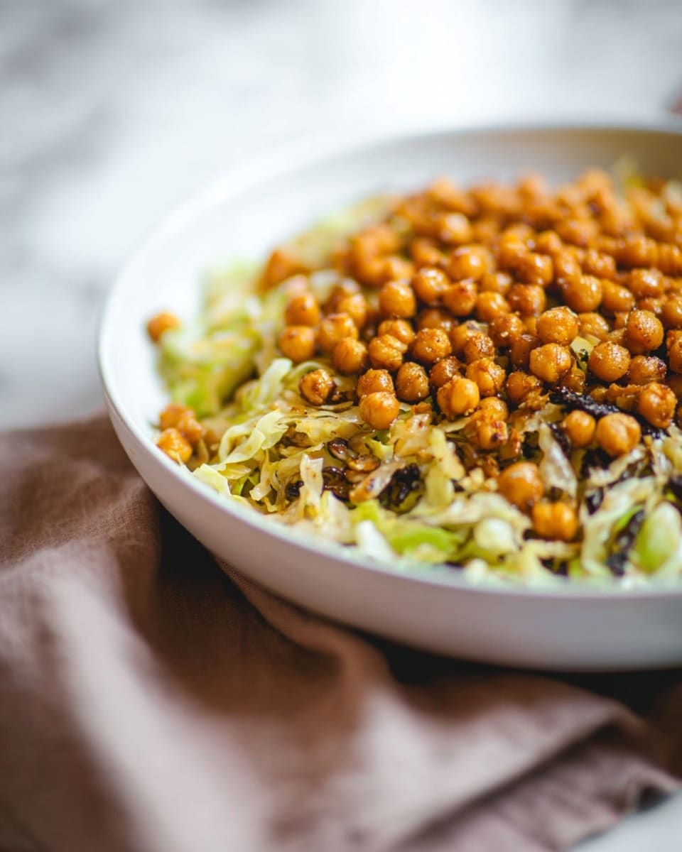 A white bowl filled with a dish that has two clear layers: the bottom layer is shredded light green cabbage mixed with some dark green and black bits, showing a mix of soft and slightly crispy textures; the top layer is round, golden-brown roasted chickpeas scattered evenly over the cabbage, adding a crunchy texture and warm color contrast. The bowl rests on a soft, light brown cloth, with a soft white marbled surface background that is blurred softly. The photo taken with an iphone --ar 4:5 --v 7