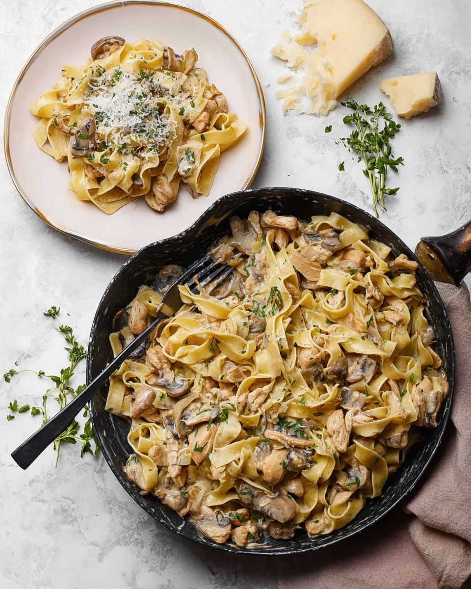 A black pan filled with wide, flat pasta ribbons mixed with light brown pieces of chicken and sliced mushrooms, all coated in a creamy light tan sauce and sprinkled with small green herbs. Next to the pan, a white plate holds a serving of the same pasta, chicken, and mushrooms layered loosely, topped with more finely chopped herbs and grated cheese that adds a light dusting of white on top. A black fork rests on the plate, partially inserted into the pasta. Around the setup, there are pieces of white cheese and green herb sprigs placed on a white marbled surface. Photo taken with an iphone --ar 4:5 --v 7