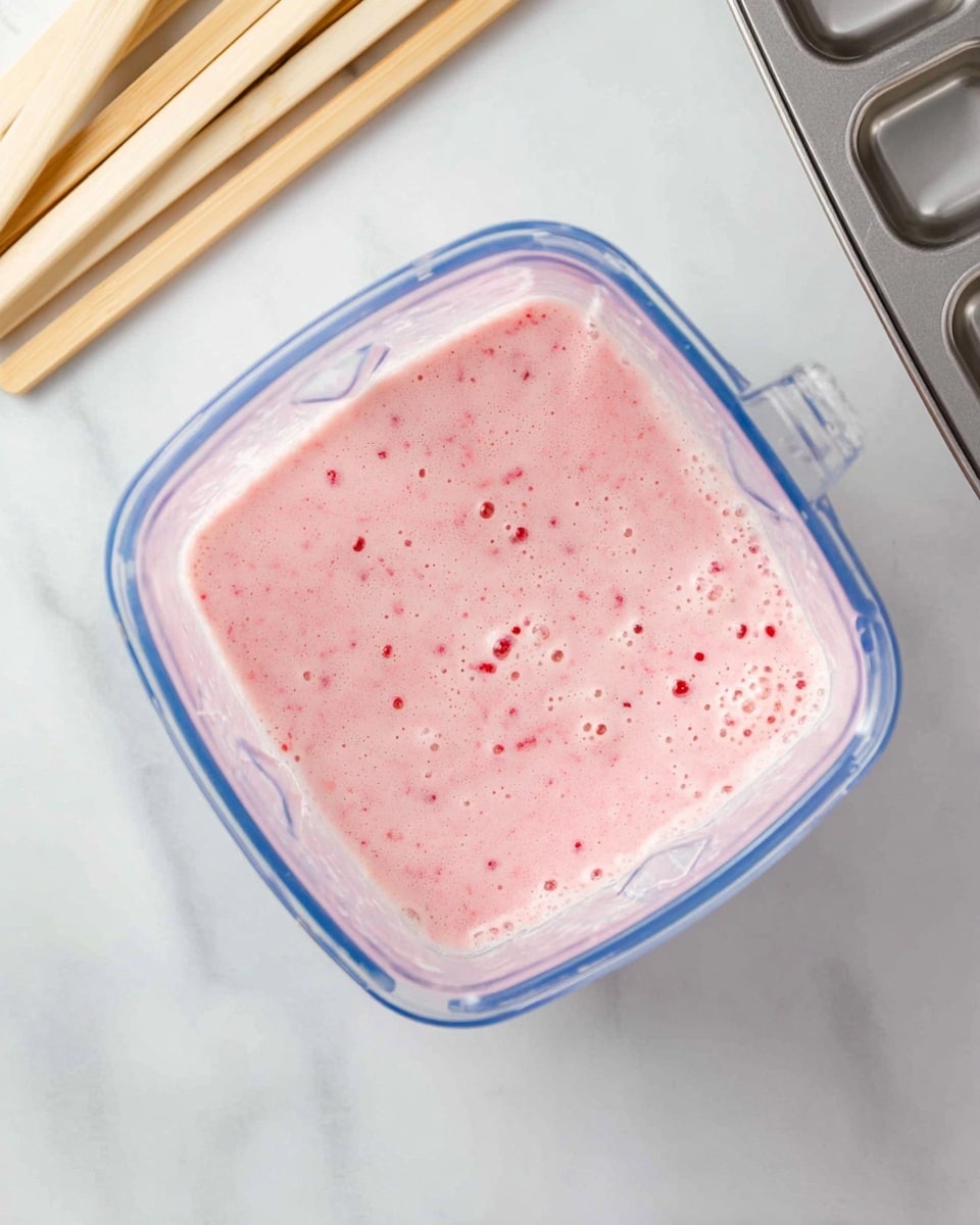 A clear blender container filled with a pink creamy mixture that has small red and green specks inside, showing tiny bubbles on the top layer. The blender container has a blue rim and is placed on a white marbled surface. To the top left are several light wooden sticks laying flat, and to the right is a metal ice cube tray. photo taken with an iphone --ar 4:5 --v 7