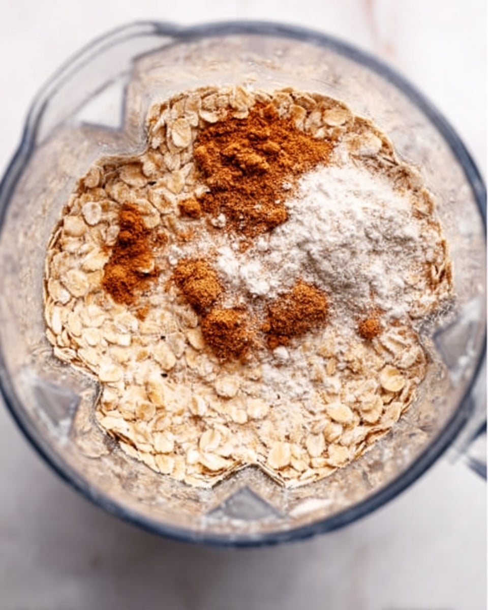 A clear blender cup seen from above, filled with dry oats covering most of the surface with a light beige color and rough texture. In the center, there are small piles of cinnamon with a rich brown color and baking powder with a soft white powdery look, sitting on top of and mixing slightly into the oats. The setting is on a white marbled surface. photo taken with an iphone --ar 4:5 --v 7