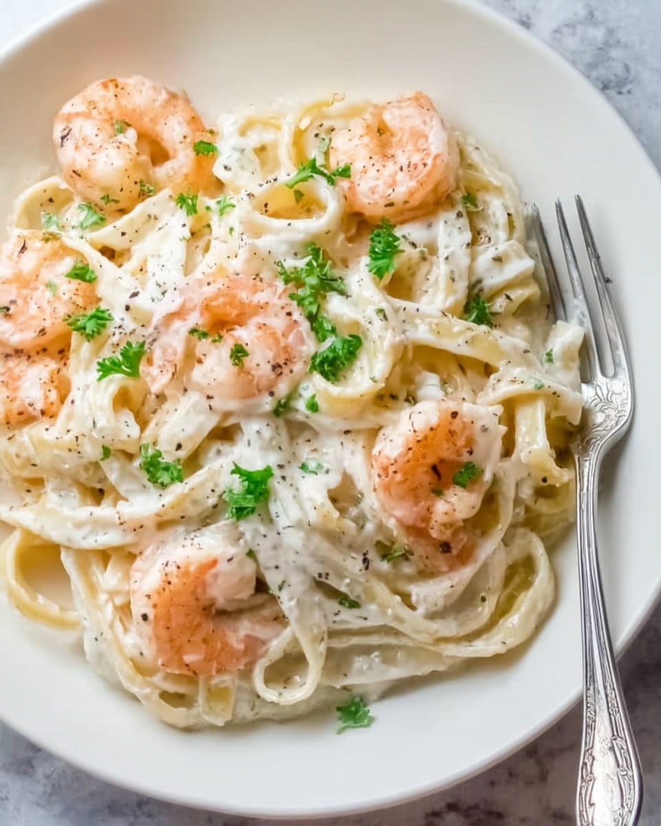 The image shows a white plate filled with creamy fettuccine pasta, covered in a smooth, white sauce with visible specks of black pepper. On top of the pasta are several pink shrimp, cooked and slightly curled, scattered evenly around the plate. Small green parsley leaves are sprinkled over the whole dish, adding a touch of color. A silver fork rests on the edge of the plate on the right side. The plate is placed on a white marbled surface. photo taken with an iphone --ar 4:5 --v 7