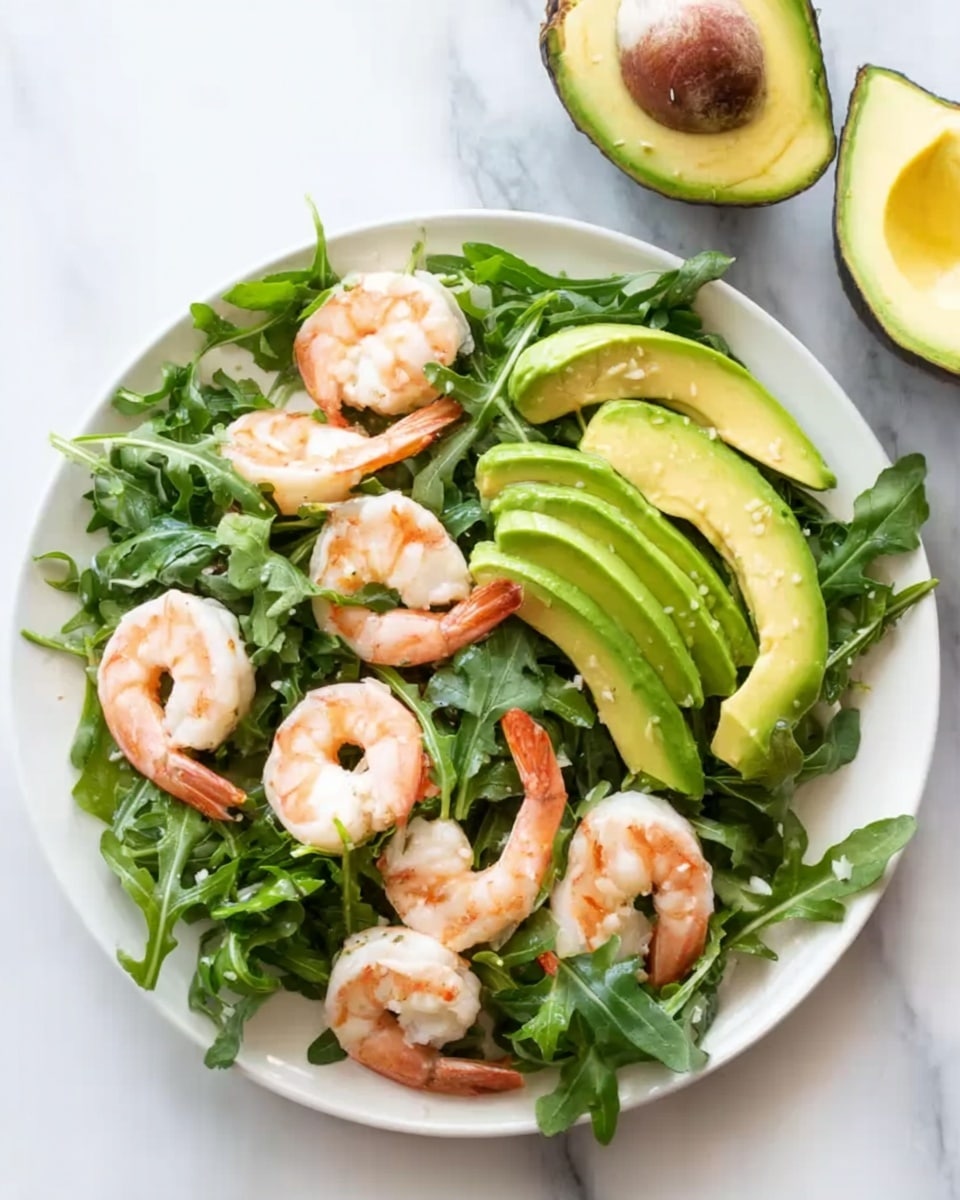 A white round plate filled with a fresh salad that has several whole cooked shrimp placed evenly over a bed of dark green leafy arugula. Slices of bright green avocado fan out neatly around the shrimp. The shrimp have a light pink and white color with a firm texture. The background is a white marbled surface with two avocado halves, one showing the seed and the other empty, positioned near the top edge of the image. Photo taken with an iphone --ar 4:5 --v 7